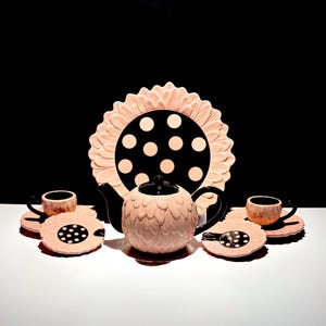 May include: A tea set featuring a teapot, two cups, and two saucers, all crafted with a light pink petal-like texture. The teapot and saucers feature a black centre with light pink polka dots, while the cups have a solid black interior.