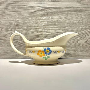 May include: A cream-colored ceramic gravy boat featuring a delicate floral pattern with blue and orange flowers, green leaves, and small yellow accents. The vessel has a curved handle, a wide pouring spout, and a scalloped base design.