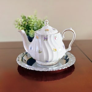 Peut inclure: Théière en porcelaine blanche avec un motif floral, posée sur un plateau argenté orné. La théière a une anse et un bec courbés, avec des bordures dorées. Le motif floral comprend des fleurs jaunes, violettes et bleues.