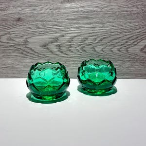 Peut inclure: Deux petits bougeoirs en verre vert vif présentant un design géométrique à facettes qui ressemble aux pétales superposés d'une fleur. Le verre est translucide et poli, créant une surface claire et réfléchissante aux bords angulaires.