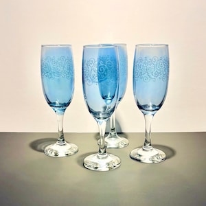 May include: Four blue champagne flutes with a decorative swirl pattern. The flutes have a clear stem and base. The glasses are arranged on a gray surface against a white background.