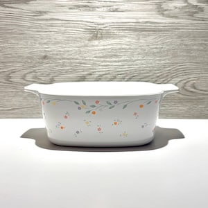 May include: A white rectangular casserole dish with integrated side handles, featuring a delicate pattern of small orange, yellow, and light purple flowers with thin green leaves and vines scattered across the exterior surface of the vessel.
