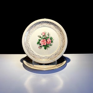 May include: Two vintage plates. The top plate is cream-coloured with a gold filigree border and a central floral design of pink roses and green leaves. The bottom plate is dark with a gold border. Both plates are on a white surface.