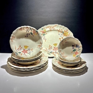 Peut inclure: Un ensemble de vaisselle en céramique crème avec des motifs floraux. L'ensemble comprend des assiettes, des bols et des soucoupes, avec un bord festonné. Le motif floral comprend des fleurs jaunes, roses et bleues avec des feuilles vertes.
