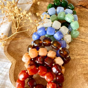 Peut inclure: Une collection de bracelets élastiques composés de perles lisses de forme carrée dans une variété de couleurs, notamment vert foncé, bleu clair, vert sauge, blanc, bleu roi, brun foncé, pêche et rouge vif, séparées par de petites entretoises dorées.