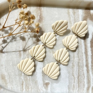 Può includere: Orecchini a forma di conchiglia color crema, con superficie strutturata e connettori color oro. Gli orecchini sono disposti in fila, mostrando il loro design a strati. Un accessorio unico.