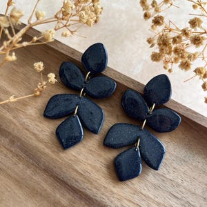 Peut inclure: Une paire de boucles d'oreilles pendantes bleu marine foncé avec un motif à plusieurs feuilles. Chaque boucle se compose de trois éléments en forme de feuille reliés par des anneaux métalliques dorés, avec une finition légèrement pailletée.