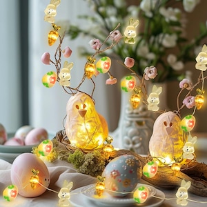 May include: Easter-themed string lights featuring bunny and carrot-shaped lights, along with illuminated egg-shaped lights. The lights are strung around decorative branches with pink flowers and two illuminated bird-shaped decorations. The scene is set on a table.