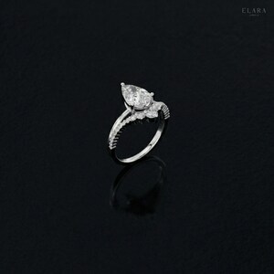 May include: A silver ring with a pear-shaped diamond center stone. The ring features a delicate band with smaller accent stones and a detailed setting. The ring is displayed on a black surface, highlighting its sparkle and design.
