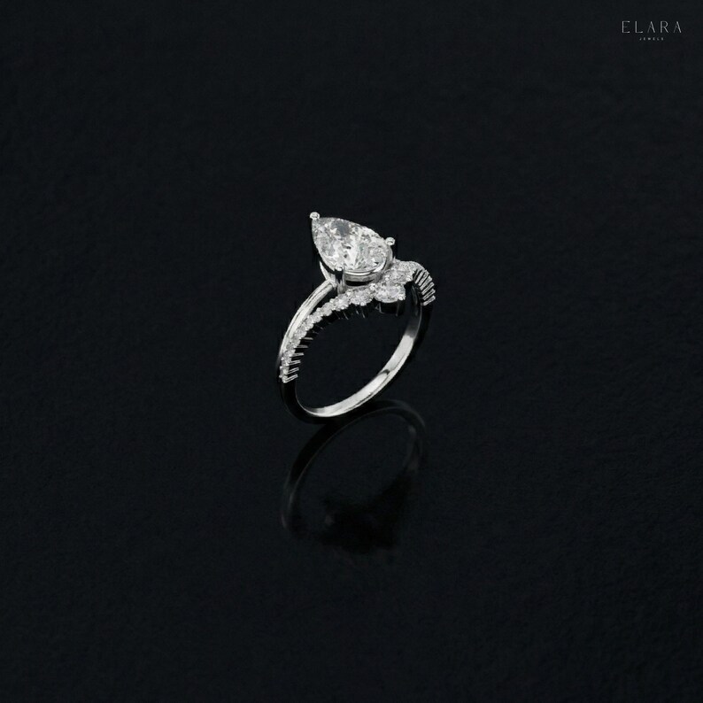 May include: A silver ring with a pear-shaped diamond center stone. The ring features a delicate band with smaller accent stones and a detailed setting. The ring is displayed on a black surface, highlighting its sparkle and design.