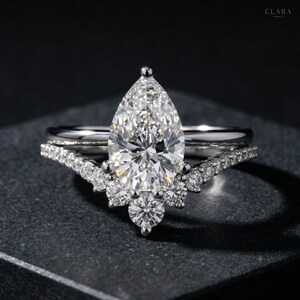 May include: A diamond engagement ring set with a pear-shaped center stone and a curved band. The ring features a sparkling, clear diamond and is set on a dark surface. The band is adorned with smaller diamonds.