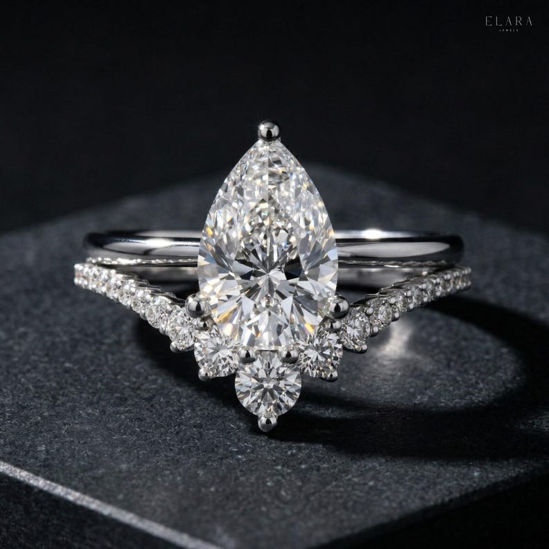 May include: A diamond engagement ring set with a pear-shaped center stone and a curved band. The ring features a sparkling, clear diamond and is set on a dark surface. The band is adorned with smaller diamonds.
