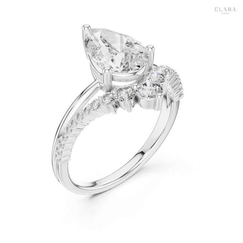 May include: A silver engagement ring with a pear-shaped diamond center stone. The band features a split shank design with smaller accent diamonds and a cluster of diamonds on the side. The ring is set against a white background.
