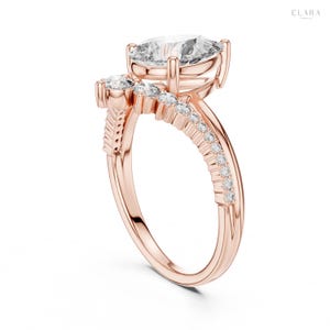 May include: Rose gold engagement ring featuring a large, clear, cushion-cut center stone. The band is adorned with smaller, sparkling accent stones and a unique side design. The ring is set against a white background, highlighting its elegant design.