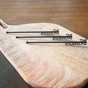 Pode incluir: Uma tábua de corte de madeira com uma pega, com três palitos pretos para servir. Cada palito tem o texto "THE ANDERSONS" em uma fonte serif. A tábua tem um grão de madeira natural.