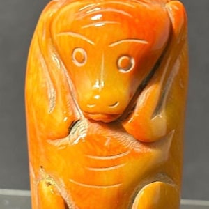 May include: A carved orange snuff bottle featuring a stylized animal figure with rounded ears, wide eyes, and a small mouth. The cylindrical body is topped with a matching orange cap, showcasing a smooth, polished surface with natural color variations.