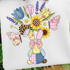 May include: White square cushion featuring a patchwork bunny holding a bouquet of flowers and butterflies. The bouquet includes sunflowers, daisies, tulips, and lavender. The bunny has a pink bow and the butterflies have pink gingham wings.