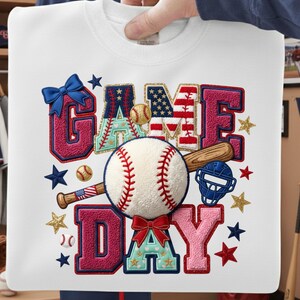 Pode incluir: Camisola branca com um design de beisebol. O design inclui as palavras "GAME DAY" em vermelho, azul e branco, juntamente com bolas de beisebol, estrelas e um bastão. A camisola é segurada por uma pessoa.