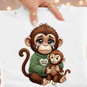 May include: White sweatshirt with a distressed illustration of a crying monkey in a green sweater, hugging a smaller monkey. The sweater has the text "Love Is Made".