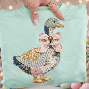 Könnte beinhalten: Hellblaues Sweatshirt mit einem aufgenähten Gänsemotiv im Patchwork-Stil. Die Gans besteht aus verschiedenen gemusterten Stoffen, darunter Blumen-, Streifen- und Vichy-Muster. Eine rosa Schleife mit Spitzenbesatz und eine Perlenknopfan der Gans Hals.