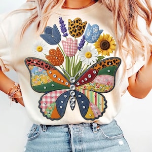 May include: Cream-coloured t-shirt featuring a colourful butterfly design with floral accents. The butterfly has patchwork wings with denim and patterned fabric. The design includes daisies, tulips, and a sunflower, embellished with rhinestones.