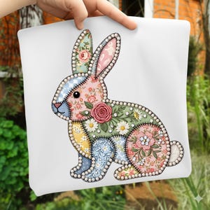 Peut inclure: Un coussin blanc avec un motif de lapin en patchwork. Le lapin est composé de divers motifs floraux roses, bleus, jaunes et verts, bordés d'une bordure perlée. Une grande rose rose est centrée sur le corps du lapin.
