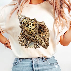 Puede incluir: Camiseta color crema con un diseño detallado de concha marina. La concha está adornada con lentejuelas doradas, pedrería y un estampado de leopardo. Adornos adicionales con temática marina incluyen un caballito de mar y pequeñas conchas.