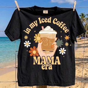 Könnte beinhalten: Schwarzes T-Shirt mit dem Text "in my Iced Coffee Mama era" in einem Retro-Schriftzug. Die Grafik zeigt eine Hand, die ein Eiskaffee-Getränk mit Schlagsahne hält, umgeben von Blumen und Sternen.