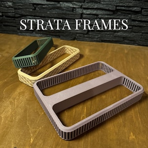 Puede incluir: Tres marcos rectangulares en verde, beige y malva, con un diseño ranurado. Los marcos están dispuestos sobre una superficie de madera, con el texto "STRATA FRAMES" en la parte superior. El logotipo de RAWForm STUDIO está en la esquina inferior izquierda.