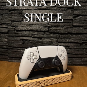 Pode incluir: Um controle de jogo branco e preto repousando em uma base de madeira com um design geométrico. O texto "STRATA DOCK SINGLE" é exibido acima do controle. A base é feita de madeira clara.