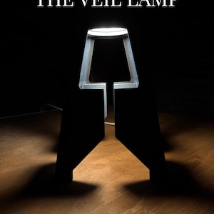 Könnte beinhalten: Eine moderne Lampe mit geometrischem Design, die ein warmes Licht ausstrahlt. Die Lampe besteht aus dunklem Material und trägt oben den Text "THE VEIL LAMP". Der Sockel ist dunkel, und das Licht scheint auf eine Holzoberfläche.