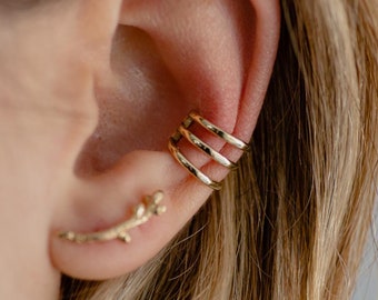 Könnte beinhalten: Nahaufnahme eines Ohrs mit zwei goldenen Ohrringen. Der Ohrring am oberen Ohr ist ein dreifacher Ohrring. Der Ohrring am unteren Ohr ist eine dünne, gebogene Stange mit einem kleinen, filigranen Design.