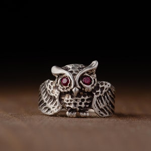 May include: A silver owl ring with red gemstone eyes. The owl is depicted with its wings spread out.