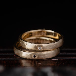 Könnte beinhalten: Zwei goldene Eheringe, die übereinander gestapelt sind. Jeder Ring hat eine strukturierte Oberfläche und einen kleinen Edelstein. Ein Ring hat einen klaren Edelstein, der andere einen dunklen Edelstein.