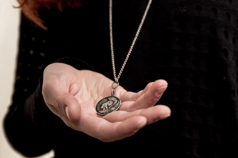 May include: A silver chain necklace with a round pendant featuring a detailed design of a creature. The pendant is held in a person's open hand.