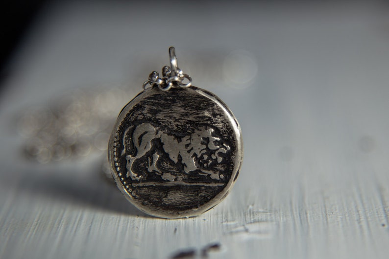 May include: A silver pendant with a detailed lion design. The pendant is round and has a textured surface.