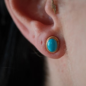 May include: A close-up of a turquoise gemstone earring with a gold setting. The earring is worn in the upper lobe of the ear.