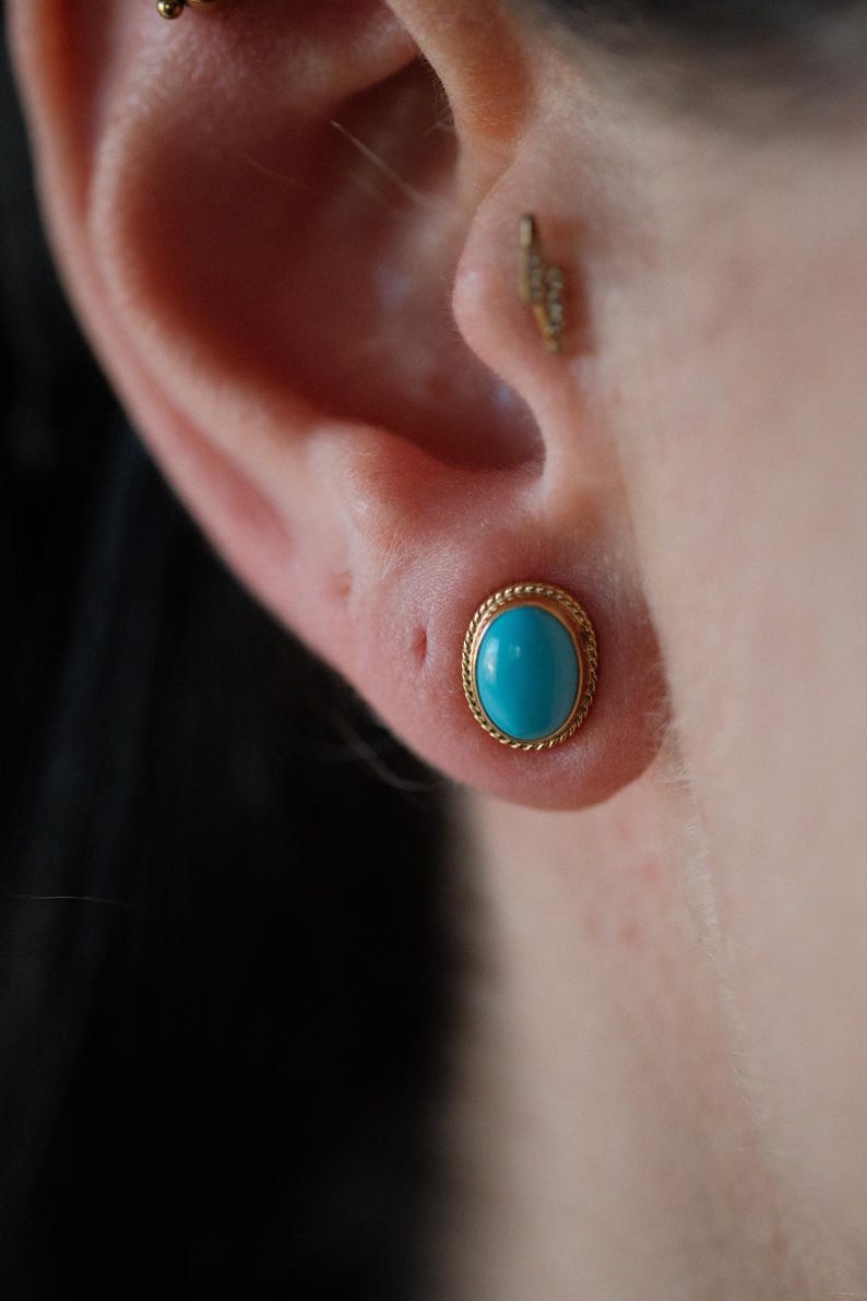 May include: A close-up of a turquoise gemstone earring with a gold setting. The earring is worn in the upper lobe of the ear.