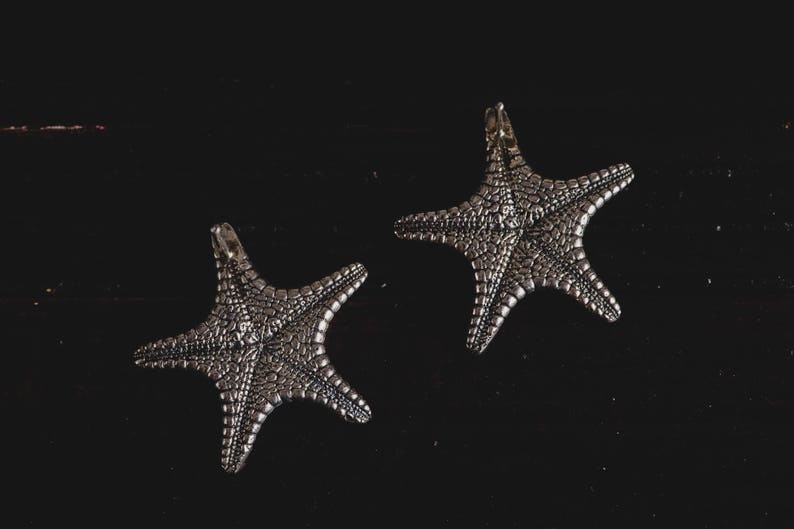 May include: Two silver starfish charms on a black background. The starfish are detailed and have a textured surface.