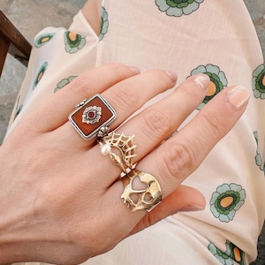 Op de afbeelding: Een hand met drie ringen. De ring op de wijsvinger is een zilveren ring met een vierkante bruine steen en een bloemmotief. De ring op de middelvinger is een zilveren ring met een spinnenwebontwerp en een parel. De ring op de ringvinger is een gouden ring met een hartvormig ontwerp.