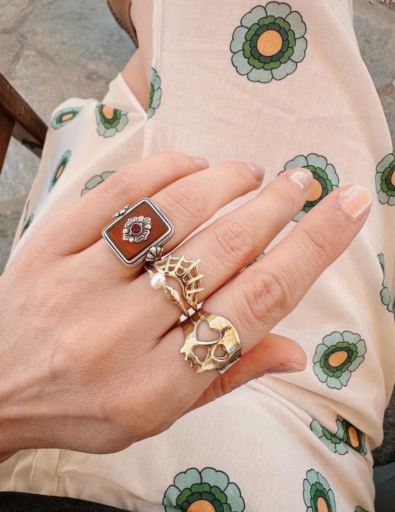 Op de afbeelding: Een hand met drie ringen. De ring op de wijsvinger is een zilveren ring met een vierkante bruine steen en een bloemmotief. De ring op de middelvinger is een zilveren ring met een spinnenwebontwerp en een parel. De ring op de ringvinger is een gouden ring met een hartvormig ontwerp.