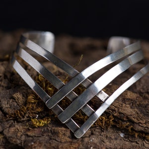 May include: A silver bracelet with a geometric design. The bracelet is made of multiple interwoven bands of metal, creating a modern and stylish look.