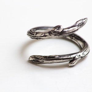 Può includere: Un anello argentato a spirale, che ricorda un ramo d'albero. L'anello presenta una superficie strutturata con incisioni dettagliate, creando un'estetica naturalistica. L'anello è posto su uno sfondo bianco.
