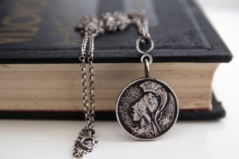 May include: A silver chain necklace with a round pendant featuring a detailed profile of a person wearing a helmet. The pendant is hanging on top of a closed book.