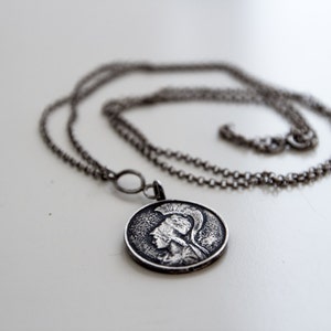 May include: A silver chain necklace with a round pendant featuring a detailed profile of a person wearing a helmet.