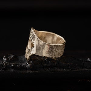 Op de afbeelding: Zilveren ring met een schedelontwerp. De ring is getextureerd en heeft een brede band.
