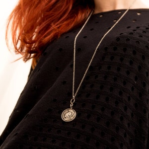 May include: A silver chain necklace with a round pendant featuring a detailed design. The necklace is worn by a person with red hair and a black, open-weave top.