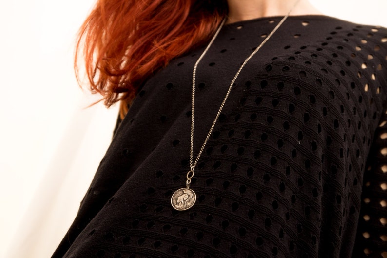 May include: A silver chain necklace with a round pendant featuring a detailed design. The necklace is worn by a person with red hair and a black, open-weave top.