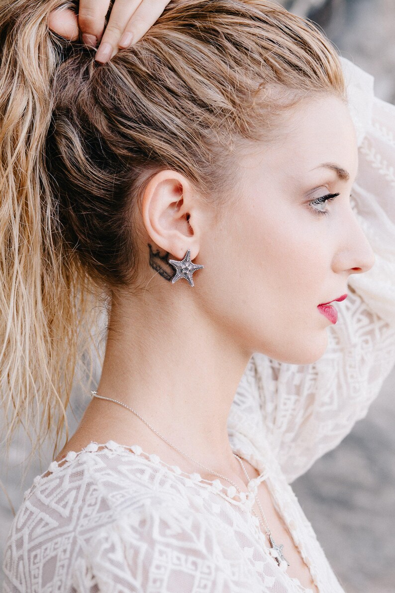 May include: A woman with blonde hair wearing a white lace top and a silver starfish earring.