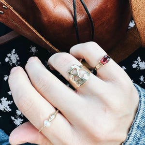 Op de afbeelding: Een hand met drie gouden ringen. De ring op de wijsvinger heeft een uitgesneden ontwerp van twee harten. De ring op de middelvinger heeft een rode edelsteen. De ring op de pink heeft een kleine parel.